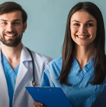 A smiling woman in blue medical scrubs holding a blue clipboard stands in front of a blurred man in a white doctor coat and stethoscope against a light blue background.